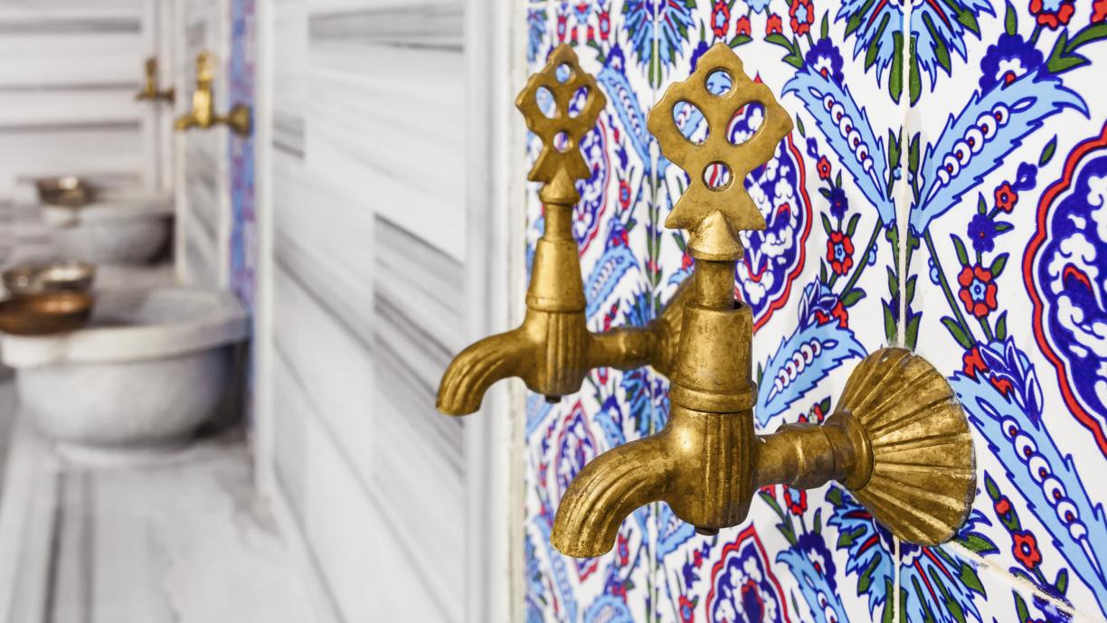 Kaksi koristeellista messinginväristä vesihanaa seinässä, joka on päällystetty värikkäillä turkkilaisilla laatoilla.