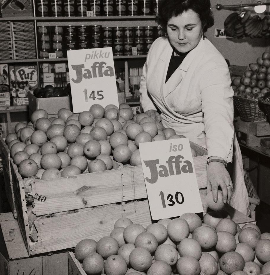 Mustavalkoinen valokuva kaupasta. Etualalla on kaksi suurta laaria täynnä appelsiineja ja niiden päällä kyltit, joissa lukee "Jaffa" ja hinnat: pikku-Jaffa 1,45 ja iso-Jaffa 1,30.