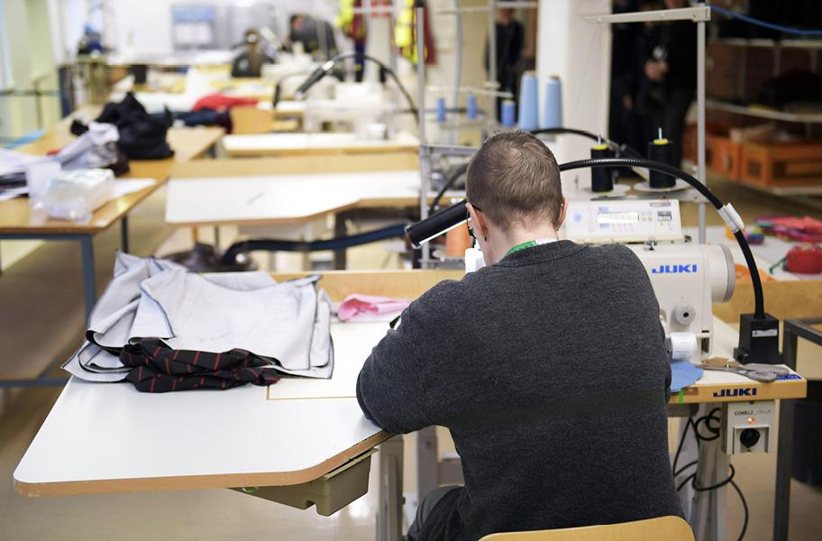 Ompelimon pöytiä, lamppuja ja ompelukoneita sekä harmaapuseroinen hahmo istumassa koneen ääressä. Pöydällä on kangaspinoja ja muita ompelutarvikkeita.