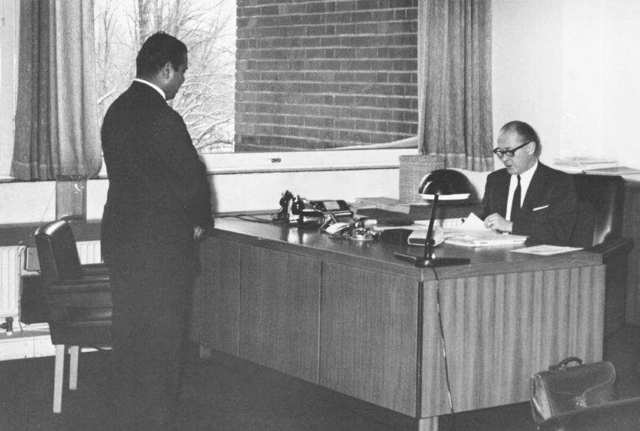 Mustavalkoinen valokuva, jossa vanhempi herra istuu työpöydän ääressä ja nuorempi mies seisoo häntä vastapäätä.