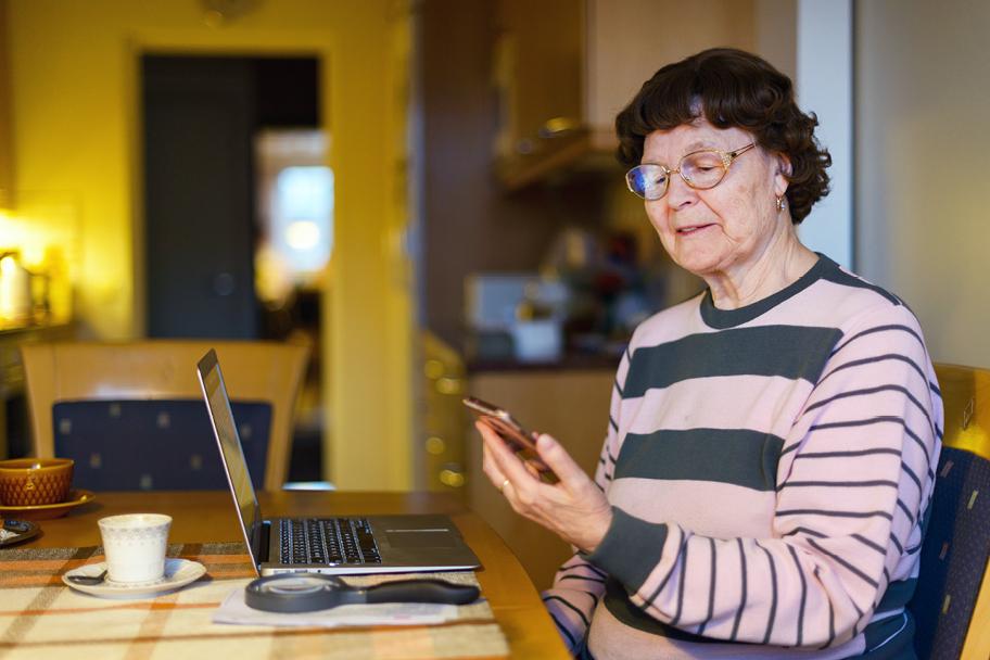 Ikääntynyt nainen istuu pöydän ääressä pidellen kädessään älypuhelimella. Pöydällä on avattu läppäri.