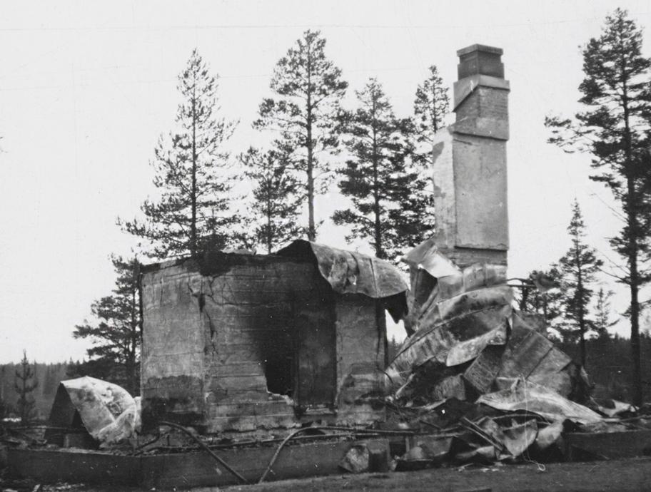 Mustavalkoinen valokuva, jossa näkyy vaurioitunut piippu, muuri ja kivijalka sekä muuta tuhoutunutta rakennusmateriaalia keskellä avaraa metsämaisemaa.