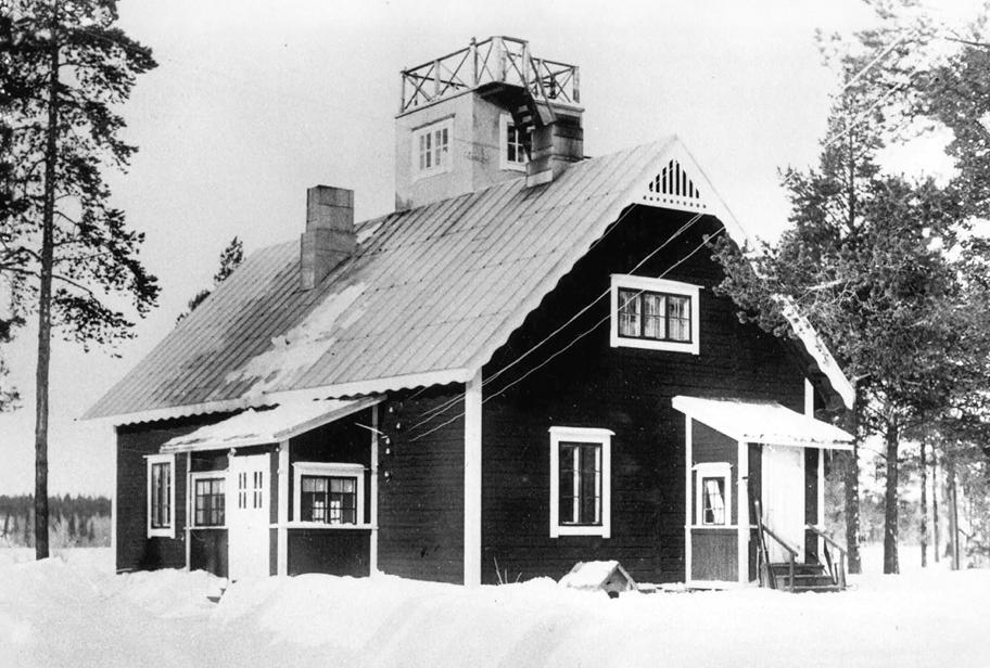 Mustavalkoinen valokuva kaksikerroksisesta harjakattoisesta puurakennuksesta lumisessa maisemassa.